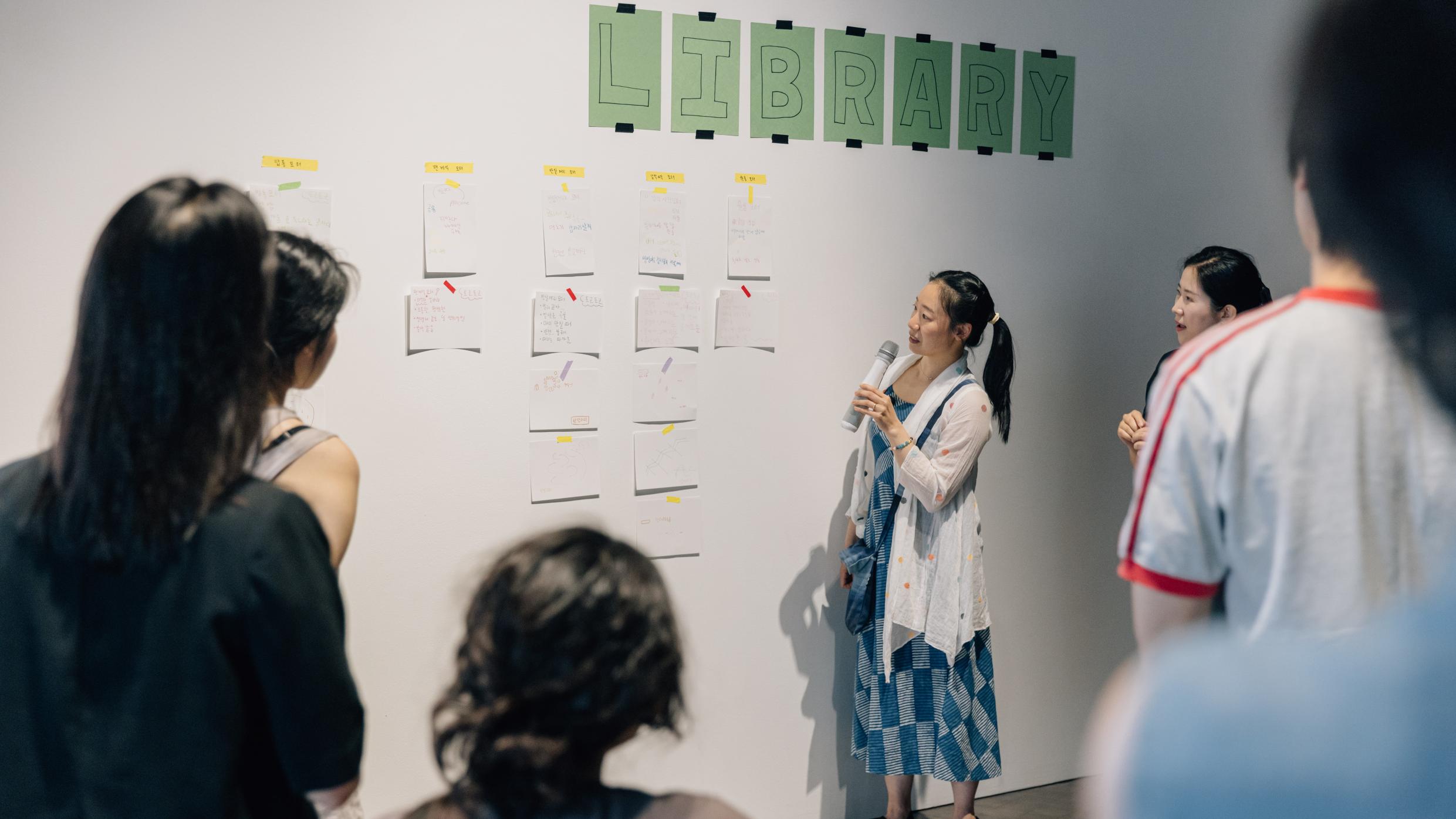 마이크를 든 발표자가 벽에 붙은 손그림 메모와 아이디어 종이들을 가리키며 설명하고 있고, 여러 사람이 둘러서서 듣고 있는 실내 워크숍 장면. 벽 위에는 “LIBRARY” 글자가 붙어 있다.